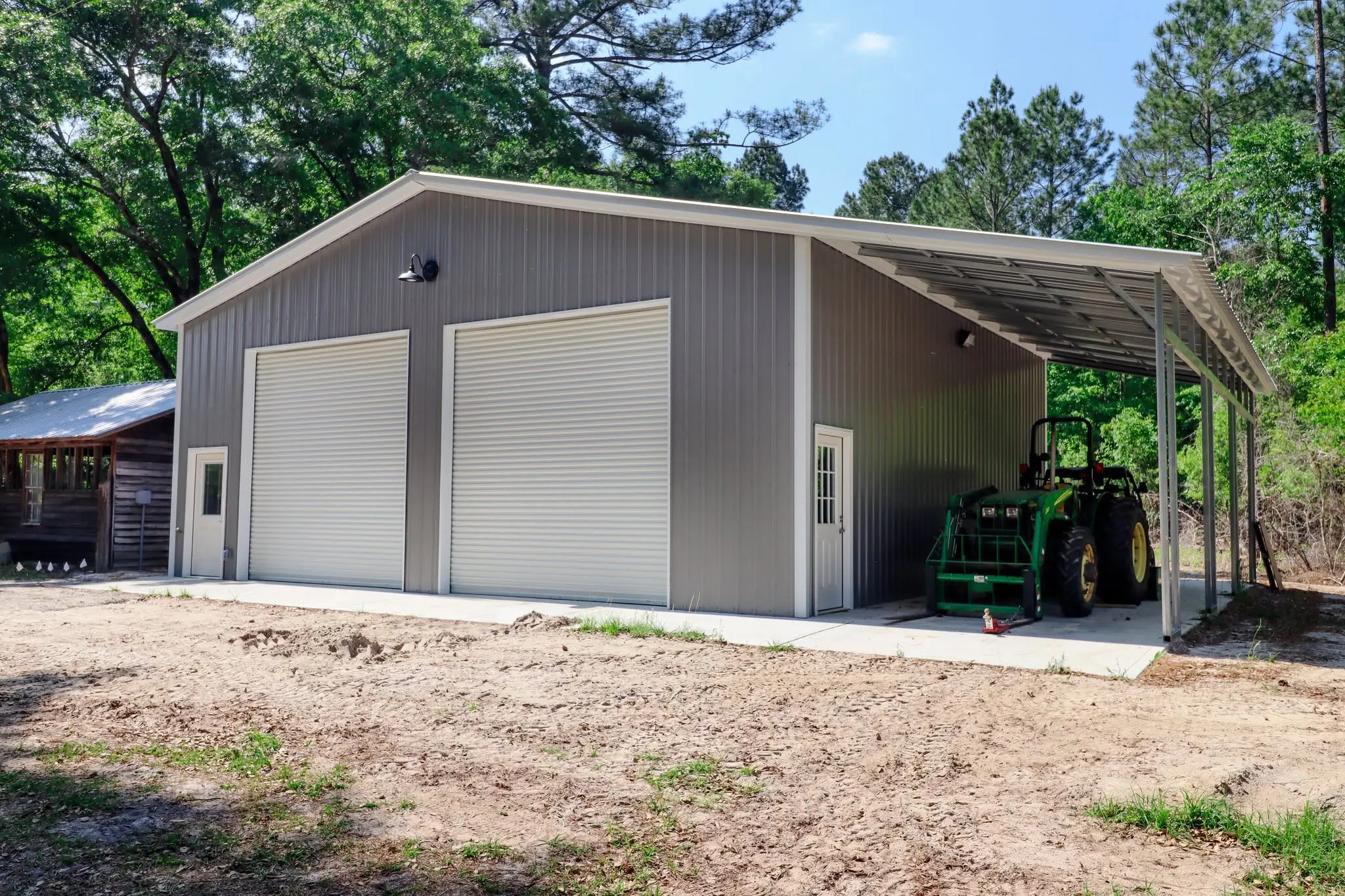 Gray steel garage with two roll-up doors and a covered side area sheltering a tractor