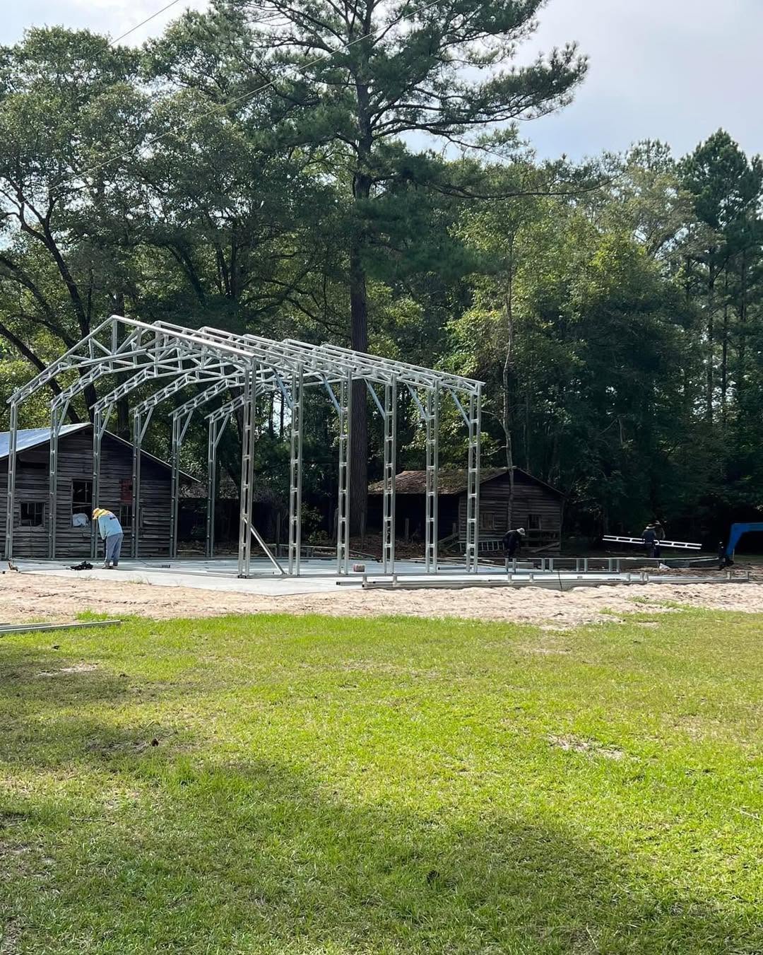 Crew erecting a steel building frame beside existing outbuildings